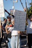 A group of people are gathered outdoors holding signs and taking photographs. One prominent sign reads a message concerning Israel and Palestine. The sky is clear, and the people are casually dressed.