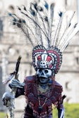 A person wearing an elaborate ceremonial costume featuring intricate designs, feathered headdress with peacock feathers, and a skull mask. The outfit includes vibrant patterns and colors, complemented by various accessories and face paint.