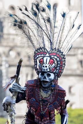 A person wearing an elaborate ceremonial costume featuring intricate designs, feathered headdress with peacock feathers, and a skull mask. The outfit includes vibrant patterns and colors, complemented by various accessories and face paint.