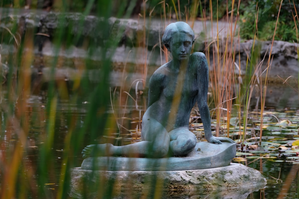 una statua di una donna seduta su una roccia in uno stagno