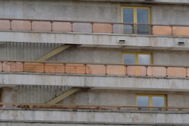 A section of a building facade featuring multiple identical balconies with orange-tinted glass railings and yellow-framed windows. The building has a concrete exterior with visible signs of weathering.