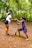 Two individuals are engaging in tree planting within a lush, green forest environment. One person is standing while holding a small tree sapling, and the other is kneeling with a shovel, preparing the ground. The area is surrounded by dense trees and foliage.