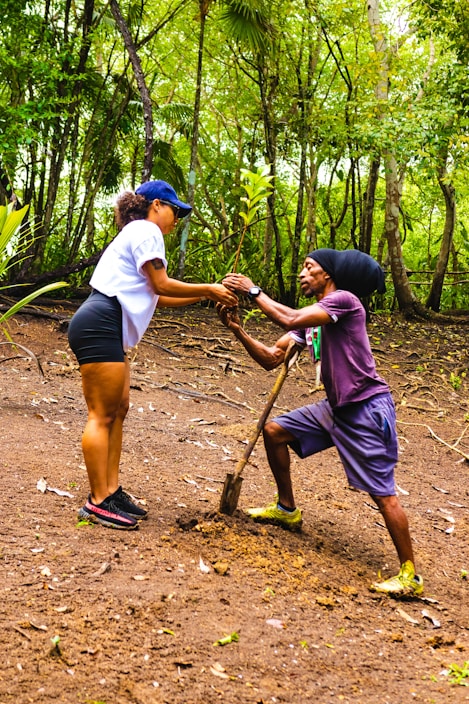 Two individuals are engaging in tree planting within a lush, green forest environment. One person is standing while holding a small tree sapling, and the other is kneeling with a shovel, preparing the ground. The area is surrounded by dense trees and foliage.