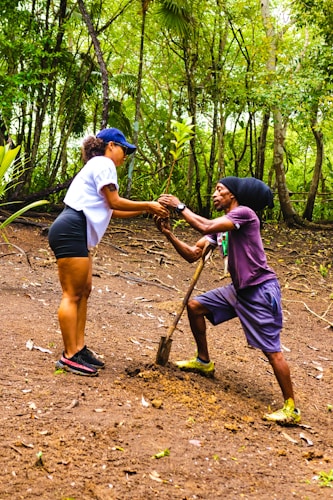 Two individuals are engaging in tree planting within a lush, green forest environment. One person is standing while holding a small tree sapling, and the other is kneeling with a shovel, preparing the ground. The area is surrounded by dense trees and foliage.