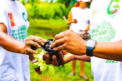 Two people are engaged in a planting activity. One person is handing a small plant or seedling to the other. Both individuals are wearing white shirts with green text, and one person is wearing a smartwatch. The hands appear to be slightly dirty, indicating involvement in gardening or planting.