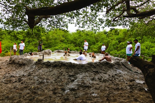 Several people are relaxing in a natural mud pool surrounded by lush green vegetation. The scene includes people both inside the muddy water and standing around the edge. Overhanging branches create a canopy overhead.