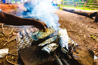 Hands lifting a foil packet of smoky maple-glazed chicken from glowing coals.