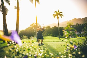 A golfer wearing a hat is in mid-swing on a sunny golf course surrounded by lush greenery and tall palm trees. The sun is setting or rising behind a hill, casting a warm glow. Wildflowers in the foreground add a touch of color to the scene.