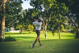 A golfer mid-swing on a sunny green fairway with tall pines in the background.