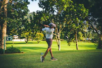 A golfer mid-swing on a sunlit green course surrounded by trees.
