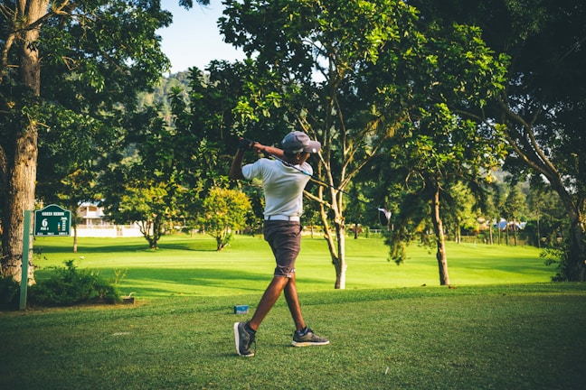 A golfer mid-swing on a sunny green fairway with tall pines in the background.