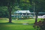 A quaint, picturesque clubhouse or country house with a green roof is nestled amidst lush greenery. Surrounding it is a well-manicured lawn and tall trees, creating a serene and inviting landscape.