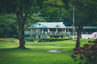 A quaint, picturesque clubhouse or country house with a green roof is nestled amidst lush greenery. Surrounding it is a well-manicured lawn and tall trees, creating a serene and inviting landscape.