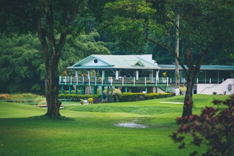 A quaint, picturesque clubhouse or country house with a green roof is nestled amidst lush greenery. Surrounding it is a well-manicured lawn and tall trees, creating a serene and inviting landscape.