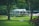 A quaint, picturesque clubhouse or country house with a green roof is nestled amidst lush greenery. Surrounding it is a well-manicured lawn and tall trees, creating a serene and inviting landscape.