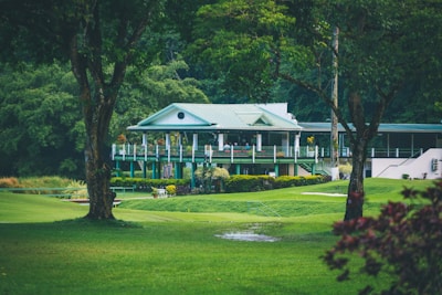 A quaint, picturesque clubhouse or country house with a green roof is nestled amidst lush greenery. Surrounding it is a well-manicured lawn and tall trees, creating a serene and inviting landscape.