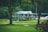 A quaint, picturesque clubhouse or country house with a green roof is nestled amidst lush greenery. Surrounding it is a well-manicured lawn and tall trees, creating a serene and inviting landscape.