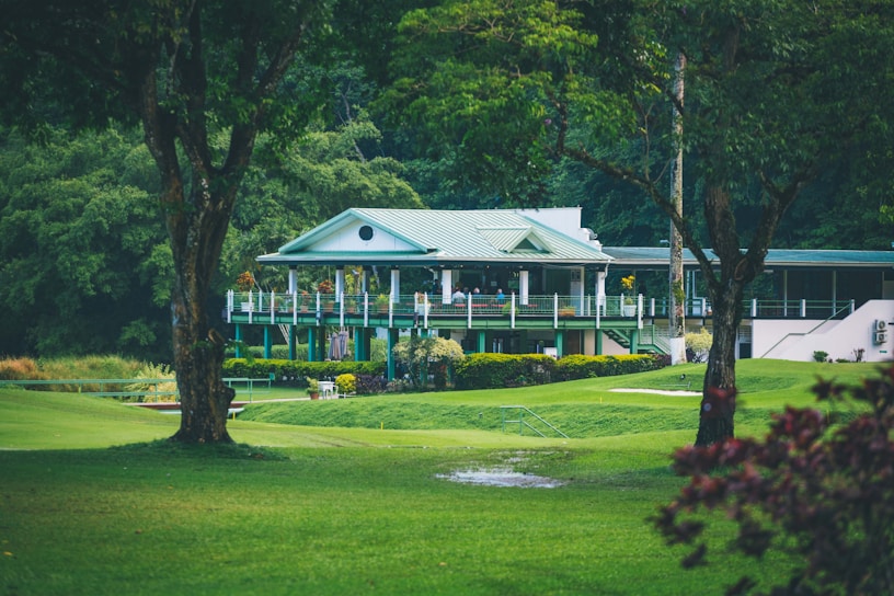 A quaint, picturesque clubhouse or country house with a green roof is nestled amidst lush greenery. Surrounding it is a well-manicured lawn and tall trees, creating a serene and inviting landscape.