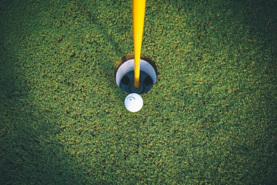 A golf ball rests near the edge of a hole on a neatly trimmed green. The yellow flagpole stands upright, indicating the location of the hole. The grass appears lush and well-maintained, providing a rich green backdrop.
