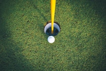 A golf ball rests near the edge of a hole on a neatly trimmed green. The yellow flagpole stands upright, indicating the location of the hole. The grass appears lush and well-maintained, providing a rich green backdrop.