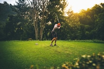 A person wearing a red cap and casual sports attire is taking a golf swing on a well-maintained grassy area surrounded by lush trees. The lighting suggests a setting sun, casting a warm glow over the scene, and a serene natural landscape is visible in the background.