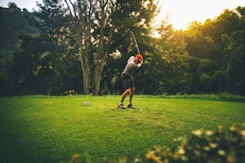 A person wearing a red cap and casual sports attire is taking a golf swing on a well-maintained grassy area surrounded by lush trees. The lighting suggests a setting sun, casting a warm glow over the scene, and a serene natural landscape is visible in the background.