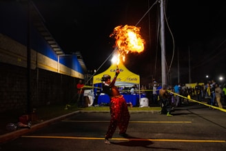 A performer is captured in mid-action during a nighttime event, skillfully breathing fire into a bright flame that illuminates the scene. The setting appears to be a street or parking area, with a yellow canopy in the background displaying the logo 'Carib'. The atmosphere is lively with a crowd of people observing the spectacle.