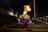 A performer is captured in mid-action during a nighttime event, skillfully breathing fire into a bright flame that illuminates the scene. The setting appears to be a street or parking area, with a yellow canopy in the background displaying the logo 'Carib'. The atmosphere is lively with a crowd of people observing the spectacle.