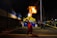 A performer is captured in mid-action during a nighttime event, skillfully breathing fire into a bright flame that illuminates the scene. The setting appears to be a street or parking area, with a yellow canopy in the background displaying the logo 'Carib'. The atmosphere is lively with a crowd of people observing the spectacle.