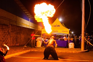 A performer is passionately engaged in a fire-breathing act at an outdoor event during nighttime. A large flame is expelled from their mouth, creating a dramatic display. People spectate from behind a roped-off area near a bright yellow tent branded with 'Carib'. The scene is vividly lit by the fire and overhead streetlights.
