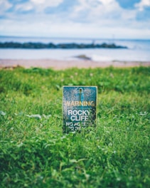 A weathered warning sign stands amidst lush green grass, cautioning about a rocky cliff. In the background, a calm sea with gentle waves and a partly cloudy sky create a serene coastal scene.