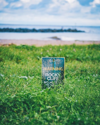 A weathered warning sign stands amidst lush green grass, cautioning about a rocky cliff. In the background, a calm sea with gentle waves and a partly cloudy sky create a serene coastal scene.