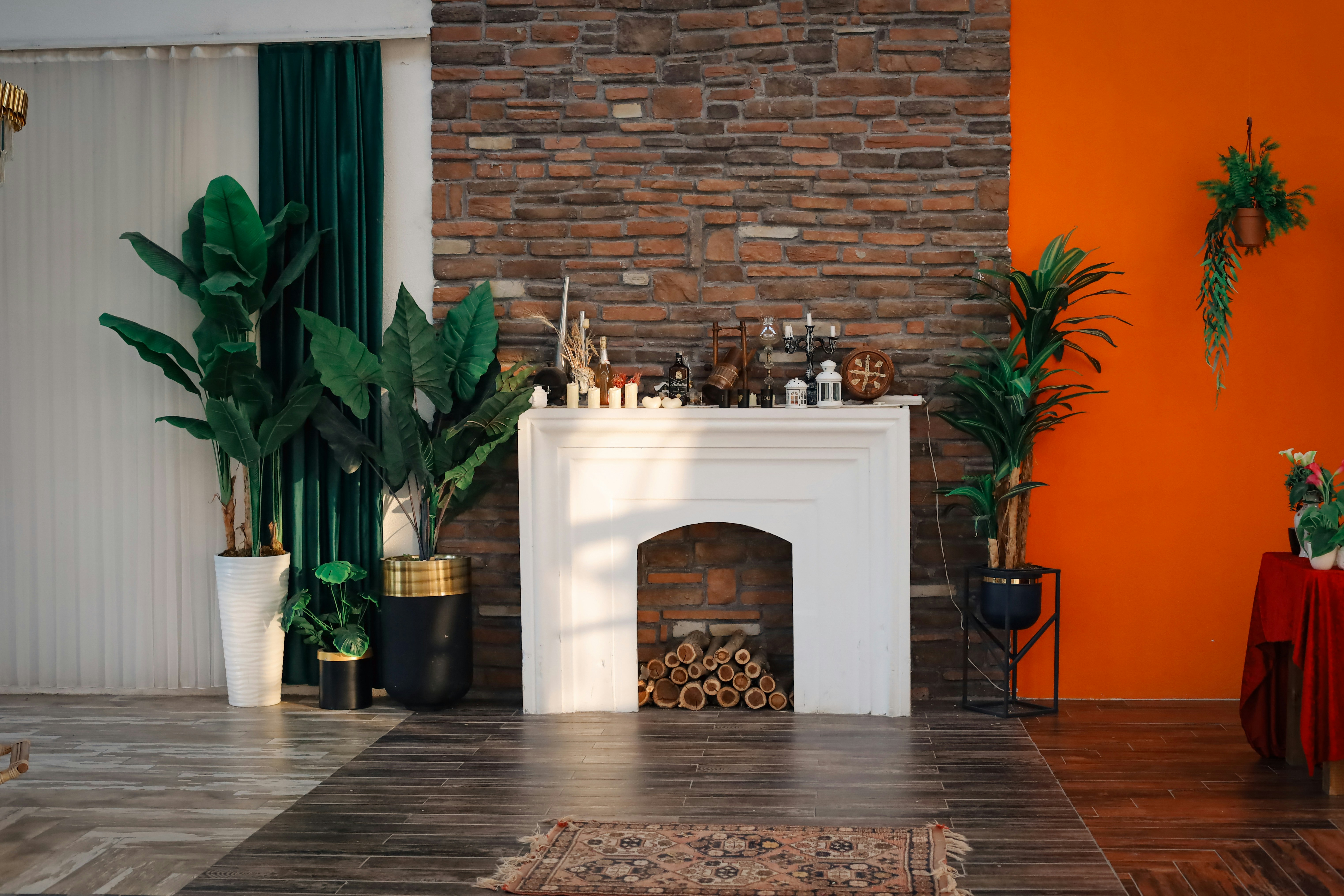 An indoor setting features a cozy fireplace set against a stone wall with several decorative plants and objects. The left side includes tall green plants in modern planters on a dark contrasting wood floor and a draped green curtain. The right side has an orange painted wall with more plants and a side table covered with a red cloth, adding a lively touch to the earthy tones.
