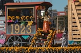 A festive cart displays a vibrant arrangement of marigold flowers and decorative papel picado, featuring the number 1923 prominently. Skeletal figures are dressed in traditional attire, creating a lively scene that suggests a cultural celebration.