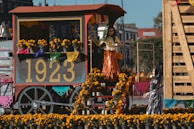 A festive cart displays a vibrant arrangement of marigold flowers and decorative papel picado, featuring the number 1923 prominently. Skeletal figures are dressed in traditional attire, creating a lively scene that suggests a cultural celebration.