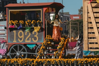 A festive cart displays a vibrant arrangement of marigold flowers and decorative papel picado, featuring the number 1923 prominently. Skeletal figures are dressed in traditional attire, creating a lively scene that suggests a cultural celebration.