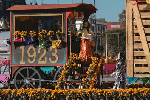 A festive cart displays a vibrant arrangement of marigold flowers and decorative papel picado, featuring the number 1923 prominently. Skeletal figures are dressed in traditional attire, creating a lively scene that suggests a cultural celebration.