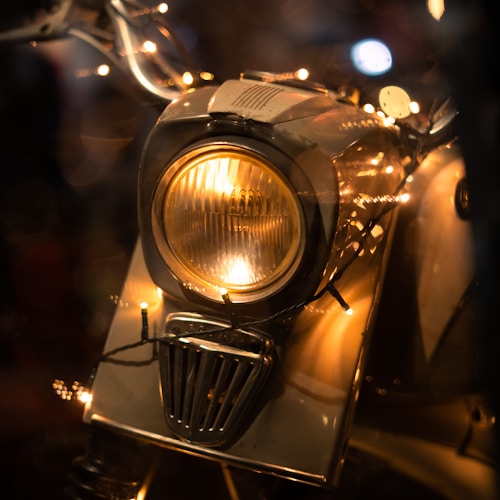 A close-up view of a vintage motorcycle headlight illuminated with warm, glowing string lights wrapped around it. The lights create a cozy, nostalgic atmosphere and highlight the metallic details of the motorcycle.