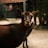 A close-up of a dark brown goat standing on a sandy surface. The goat gazes directly at the camera with a calm demeanor. Another goat is partially visible in the background, standing in front of a rustic wooden building with red and white accents.