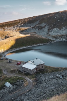 A rustic cabin with a white roof is nestled beside a serene mountain lake. The area around is rocky and sparse, with a gravel path leading to the building. Two vehicles are parked nearby, and the surrounding hills are covered with a mix of grass and scattered rocks. The sunlight casts a warm glow, creating long shadows.