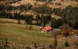 A tranquil countryside scene featuring a rustic wooden house with a red roof, surrounded by lush green fields and dense forests. In the background, rolling hills are covered with a mix of evergreen and deciduous trees. A wooden fence runs along the foreground, enhancing the pastoral ambiance.