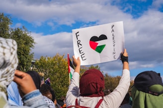 A person is holding up a sign with the words 'Free Palestine' written on it, featuring a heart filled with the colors of the Palestinian flag. The scene includes several people in the foreground, with a backdrop of trees and a partly cloudy blue sky.