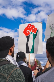 A group of people are gathered, holding signs during a protest. One prominent sign features a stylized fist colored in red, black, and green with letters that spell 'AZA'. The people are dressed in casual attire, some wearing caps and scarves. The sky is bright and partly cloudy, indicating a clear day.