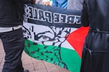 A flag featuring a stylized face with eyes visible behind a patterned scarf is held by two people. The flag includes colors black, white, green, and red, along with text. The scene is set on a tiled ground surface.