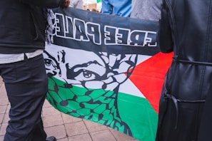 A flag featuring a stylized face with eyes visible behind a patterned scarf is held by two people. The flag includes colors black, white, green, and red, along with text. The scene is set on a tiled ground surface.