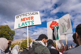 A group of people gathered outside holding protest signs. One sign reads 'Oppression Never Lasts' with the words in black and red. Another sign shows artistic illustrations with prominent red letters. The crowd appears engaged, with some individuals capturing the moment on their phones. The sky is partly cloudy, and trees are visible in the background.