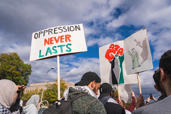 A group of people gathered outside holding protest signs. One sign reads 'Oppression Never Lasts' with the words in black and red. Another sign shows artistic illustrations with prominent red letters. The crowd appears engaged, with some individuals capturing the moment on their phones. The sky is partly cloudy, and trees are visible in the background.