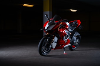 A sleek red and black motorcycle is parked in a dimly lit garage. The motorcycle has a modern, aerodynamic design with sharp lines and a glossy finish. Specific details such as the tires, handlebars, and mirrors are clearly visible. The lighting highlights the curves and details of the motorcycle while the background remains mostly dark.