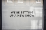 A storefront window features large, bold black text on a white background reading 'WE’RE SETTING UP A NEW SHOW'. The backdrop includes a reflection of brick walls and windows.