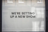 A storefront window features large, bold black text on a white background reading 'WE’RE SETTING UP A NEW SHOW'. The backdrop includes a reflection of brick walls and windows.